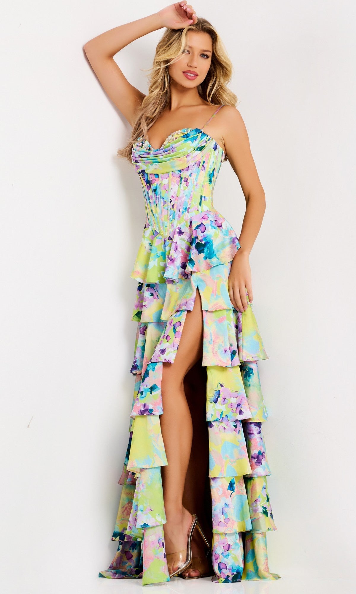 A woman models the Jovani 47560 Long Prom Dress, featuring a strapless corset bodice, watercolor print in pastel hues, tiered ruffle skirt with a high slit, and clear high heels against a white background.
